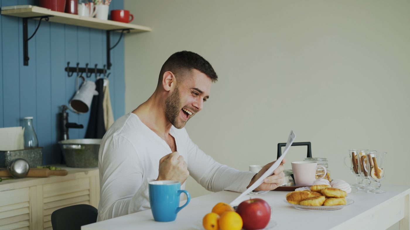 Mann am Küchentisch lacht, ballt die Faust und liest ein Schreiben, daneben Kaffeebecher und Obst.