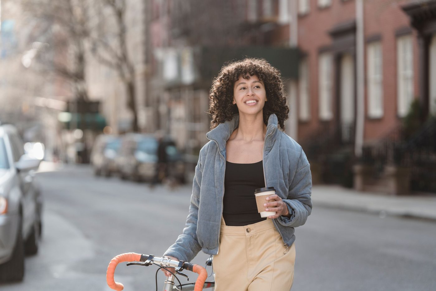 Frau schiebt ihr Fahrrad mit einem Kaffeebecher in der Hand durch eine sonnige Straße.