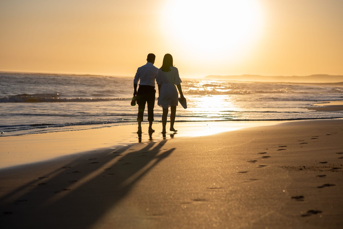 Paar läuft bei Sonnenuntergang barfuß den Strand entlang, die Schuhe in der Hand.